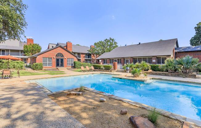 A serene outdoor pool area surrounded by lush greenery and landscaped gardens. Lounge chairs are positioned by the poolside, and a few umbrellas provide shade. Nearby, there are adjacent buildings with a brick faÃ§ade, creating a welcoming atmosphere for relaxation and leisure.