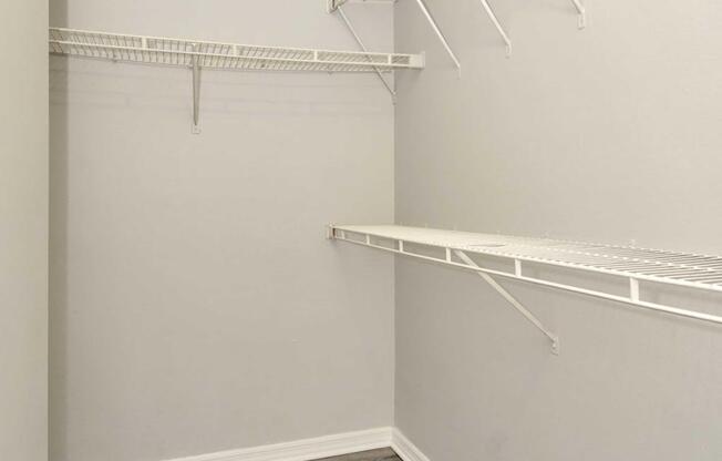 Empty closet with gray walls and white wire shelving. The shelving includes two horizontal shelves at different heights, designed for storage. The floor is a dark wood finish, adding contrast to the light-colored walls.