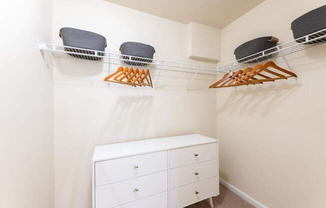 A white closet with a white dresser and a shelf with hangers and baskets.