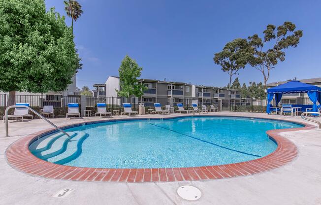 A clear swimming pool surrounded by lounge chairs under a sunny sky. Green trees are visible in the background, along with apartment buildings. A blue cabana is located near the pool, creating a relaxing outdoor space.