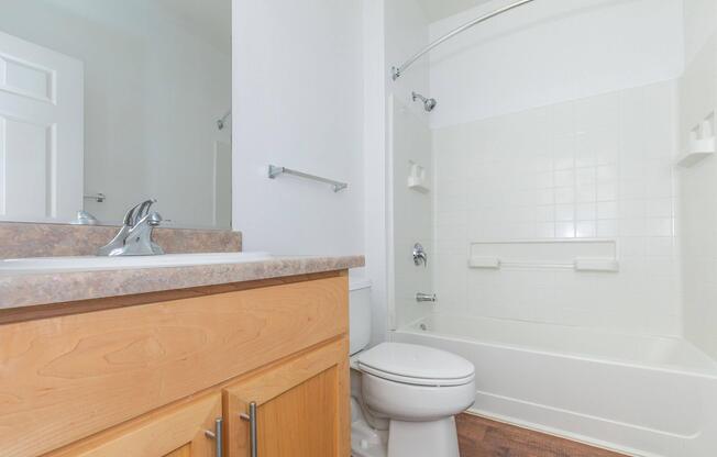 A clean and simple bathroom featuring a bathtub and shower combination, a toilet, and a wooden vanity with a sink. The walls are white, and the floor appears to be wood or laminate. There are minimal decorations, creating a tidy and modern aesthetic.