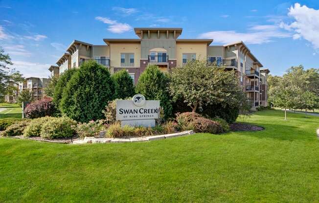 A welcoming exterior monument sign here at Swan Creek surrounded by lush green landscaping, mature trees, and manicured lawn, with the residential building rising behind for a polished and inviting community entrance.