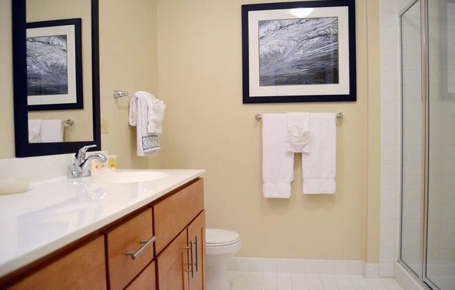 A bathroom with a spacious sink vanity, a mirror, a toilet, tile flooring, and glass doors leading to a stand-up shower.