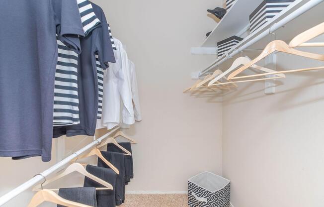 A neatly organized closet featuring various clothing items on hangers, including a striped shirt and a white blouse. The closet has a light-colored wall, a shelf with black and white storage boxes, and a decorative storage bin on the floor.