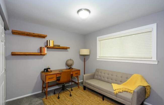A room with a grey couch, a desk with a chair, a lamp, and a window with blinds.
