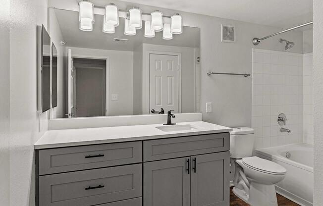 A modern bathroom featuring a double sink vanity with gray cabinets, a large mirror, and stylish light fixtures. There is a white bathtub and shower combination, a toilet, and a door leading to another room. The walls are painted light gray, creating a clean and contemporary look.