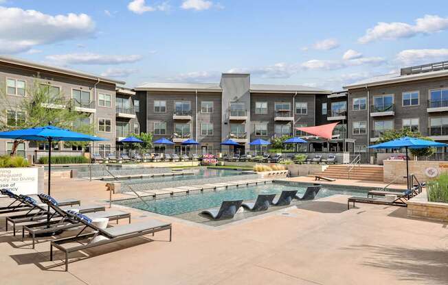 A swimming pool area with sun loungers and umbrellas in front of apartment buildings.