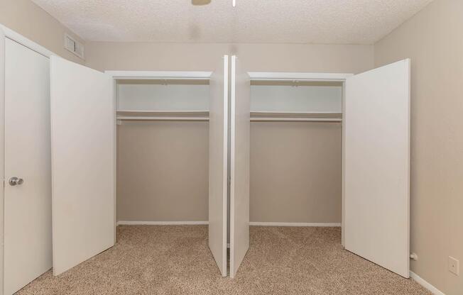 A well-lit room featuring two open white sliding closet doors, showcasing empty shelves and hanging rods. The walls are painted a light neutral color, and the floor is covered with beige carpet, creating a clean and spacious appearance.