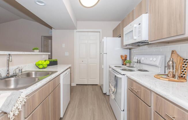 Modern kitchen featuring light wood cabinetry, white appliances, and a white countertop. The space includes a double sink, a microwave, and an oven, with a door leading to a pantry. A bowl of green apples sits on the counter, adding a pop of color to the neutral decor.