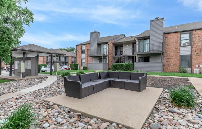 A modern outdoor seating area featuring a large, dark wicker couch surrounded by decorative stones. In the background, there are two multi-unit apartment buildings with a grassy area and a pathway leading to the entrance. The sky is clear, indicating a bright day.