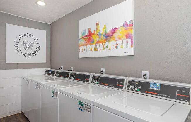 A modern laundry facility featuring several commercial washing machines lined up against a wall. To the left, there's a decorative wall art piece with the text "Laundry is an Adventure" and on the right, a colorful artwork depicting the skyline of Fort Worth. The walls are painted in neutral tones.