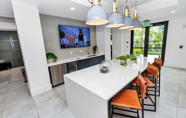 a kitchen with a large white island with orange chairs and a large tv on the wall