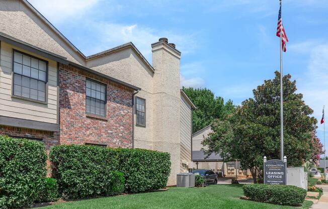 Exterior view of an apartment leasing office with a brick and stucco facade. A large American flag is displayed on a pole, surrounded by well-maintained shrubs and grass. In the background, additional buildings and trees can be seen under a clear blue sky.