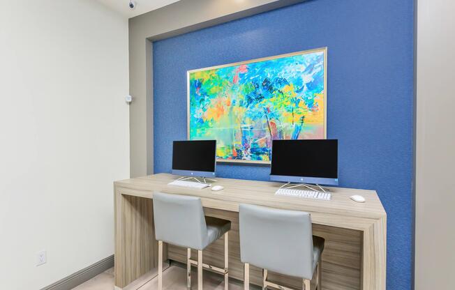 A modern workspace featuring a light wood desk with two computer monitors, each with a keyboard and mouse. The wall behind is painted a vibrant blue and adorned with a colorful abstract painting. Two stylish white chairs are placed in front of the desk, creating a clean and inviting atmosphere.