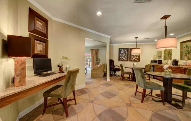 A welcoming community lounge here at Trinity Place with a built-in computer desk and chair along one wall. Round dining tables with upholstered seating sit beneath modern pendant lights, while framed wall art and soft carpeting add warmth. An open layout connects to adjacent seating areas, creating a comfortable space for work, dining, and socializing.