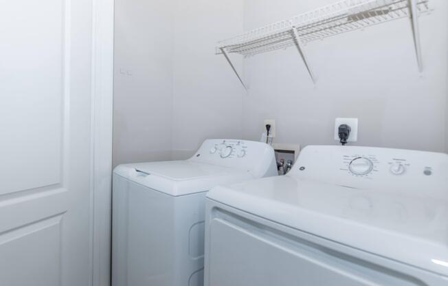 A bright laundry room featuring a stacked washer and dryer side by side. The walls are a light color, and there is a wire shelf above the appliances for additional storage. The room has a door that is partially closed, creating a cozy, functional space for laundry tasks.