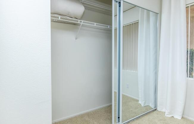 A spacious closet with mirrored sliding doors, featuring a shelf for storage and a neatly arranged interior. The closet sits beside a window dressed with white curtains, and the room has light-colored carpet and neutral walls, creating a bright and airy atmosphere.