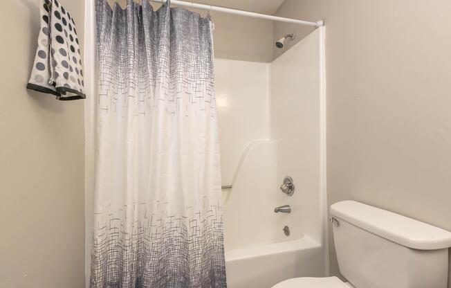 A bathroom featuring a bathtub with a shower, a white shower curtain with a gradient design, a toilet, and a towel hanging on the wall. The walls are a light color, creating a clean and simple look.