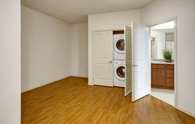 A flexible interior space here at Heritage Hills featuring durable wood-style flooring, soft neutral walls, and a recessed ceiling. The room includes a convenient in-home laundry closet with a stacked washer and dryer behind double doors, and an adjacent bathroom with a wood-tone vanity, framed mirror, and bright overhead lighting, offering versatile everyday functionality.