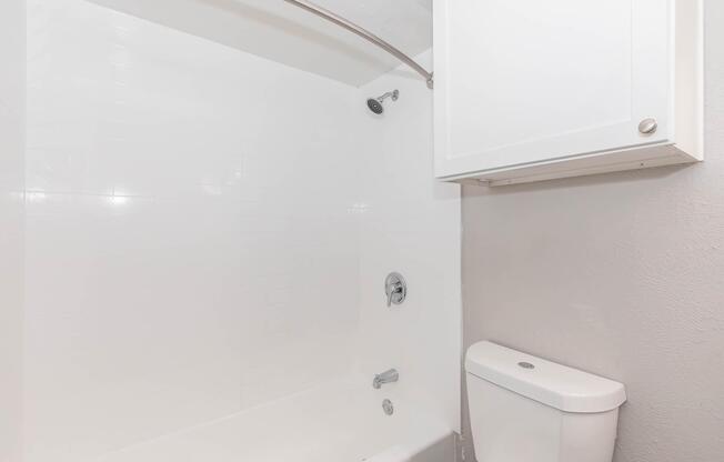 A clean and modern bathroom featuring a white bathtub with a showerhead and a curved shower curtain rod. There is a white toilet next to the tub, and a white cabinet is mounted above the toilet for storage. The walls are light-colored, creating a bright and airy atmosphere.