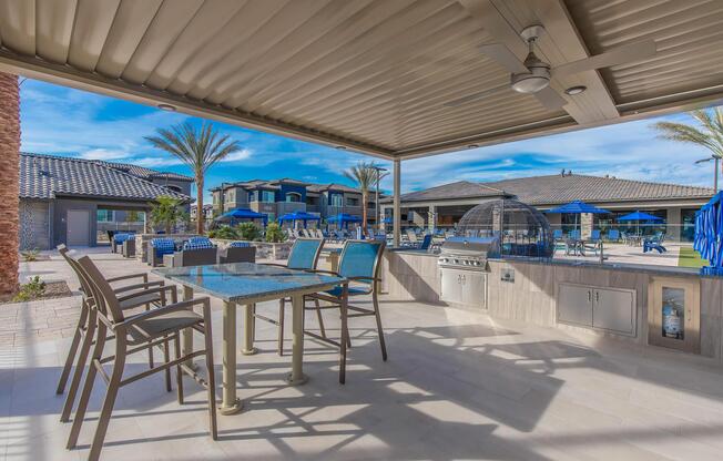 A covered outdoor dining area featuring a table with chairs, a grill, and a palm tree in the background. The scene includes a swimming pool area with blue lounge chairs and a modern apartment complex in the distance under a clear blue sky.