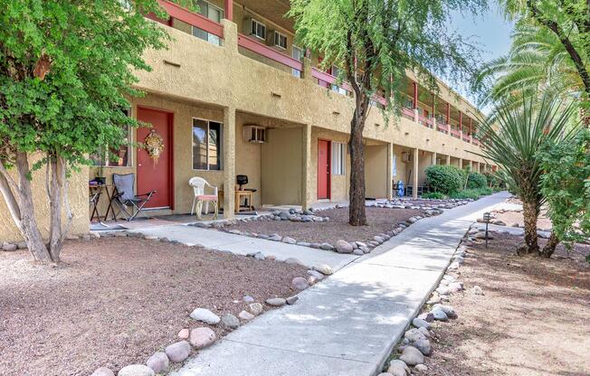 View of a walkway lined with rocks leading to a series of residential units in an apartment complex. Each unit features a front door, with a small seating area and decorative elements. Lush greenery including bushes and palm trees surrounds the area, enhancing the outdoor ambiance.