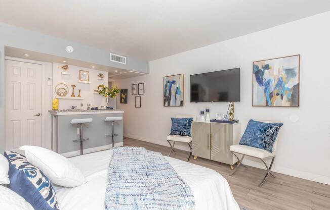 A cozy bedroom featuring a bed with white bedding and a blue throw blanket. There are two elegant chairs and a small bar area with decorative items. A flat-screen TV is mounted on the wall, and two abstract art pieces hang beside it. The room has a modern, inviting atmosphere with neutral tones and blue accents.