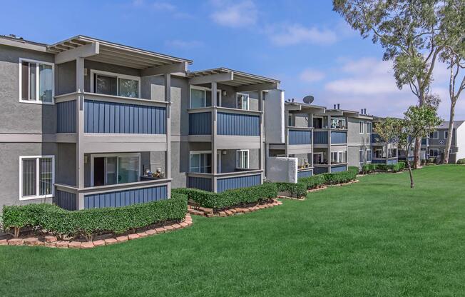 Row of modern apartment buildings with blue and gray exteriors, featuring balconies and well-maintained green lawns in front. The sky is partly cloudy, adding a serene atmosphere to the residential area.