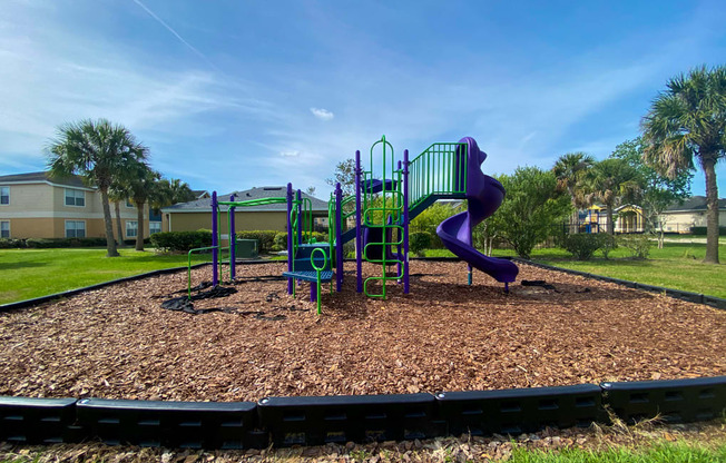 Green and blue Playground set with monkey bars in a bed of mulch with buildings and palm tree in the background