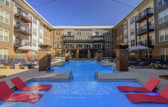 A swimming pool surrounded by red lounge chairs in front of apartment buildings.
