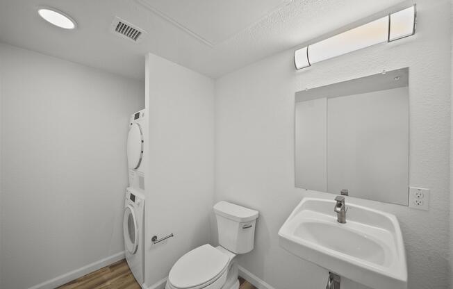 A clean and modern bathroom featuring a toilet, a small sink with a faucet, a mirror, and a stacked washer and dryer unit. The walls are painted white, and the flooring is light wood. A ceiling light fixture provides illumination in the room.