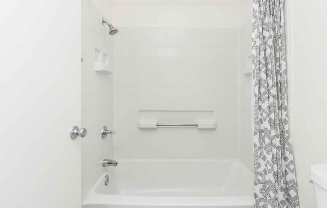 A clean and simply designed bathroom featuring a white bathtub with a showerhead above, a white tiled wall, and a patterned shower curtain. The space includes built-in shelves and a grab bar for safety, with a white door partially visible on the left side.