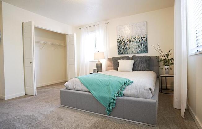 A bedroom with a bed, a grey headboard, a white bedspread, a green throw, a white pillow, a grey armchair, a white wall, a white door, a white curtain, a white lamp,