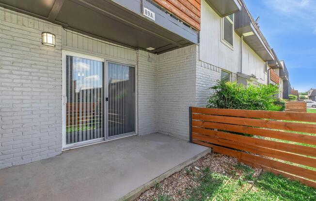 A ground-level patio area with a sliding glass door, featuring a brick wall and wooden fence. The space is surrounded by greenery and has a concrete floor. The building has a modern design with a combination of textures and colors.