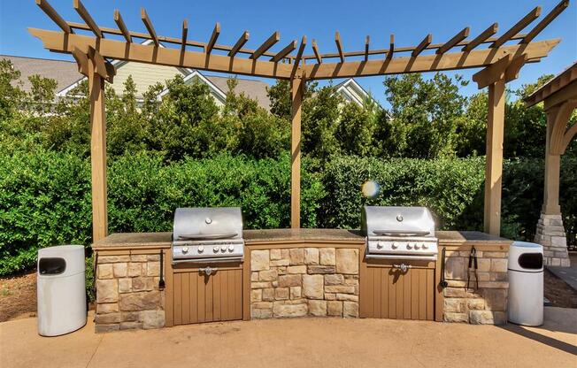 A large stone grilling station with two outdoor grills under a wooden pergola near trees