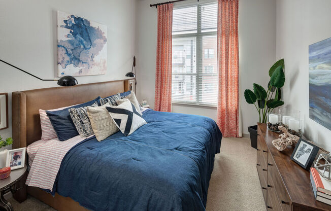 A bedroom with a blue bedspread and pillows, a wooden headboard, and a window with orange curtains.