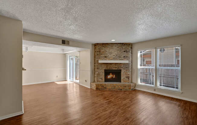 A spacious inviting living room here at Summertree Place featuring wood-style flooring, a central brick fireplace with a raised hearth, neutral walls, a textured ceiling, and large windows with blinds that fill the room with natural light while connecting the space to an adjoining area with patio access.