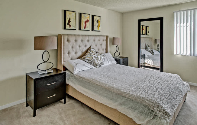 A serene bedroom here at Andorra featuring a plush upholstered bed with textured neutral bedding and accent pillows, matching nightstands with modern table lamps, soft carpet flooring, and framed artwork above the headboard, with a full-length mirror and window blinds that bring in natural light and create a calm, comfortable retreat.