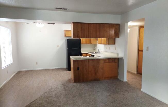 A view of a kitchen and living area in an apartment. The kitchen features wooden cabinetry, a black refrigerator, and a countertop. Natural light streams in through a window with blinds. The flooring is a mix of carpet and laminate. The space is clean and uncluttered, offering a cozy atmosphere.