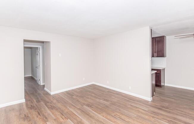 A spacious, empty room with light-colored walls and wooden laminate flooring. To the left, a doorway leads to another room. On the right, a small section of a kitchen with dark cabinets is visible. The overall atmosphere is bright and inviting, suitable for various design possibilities.