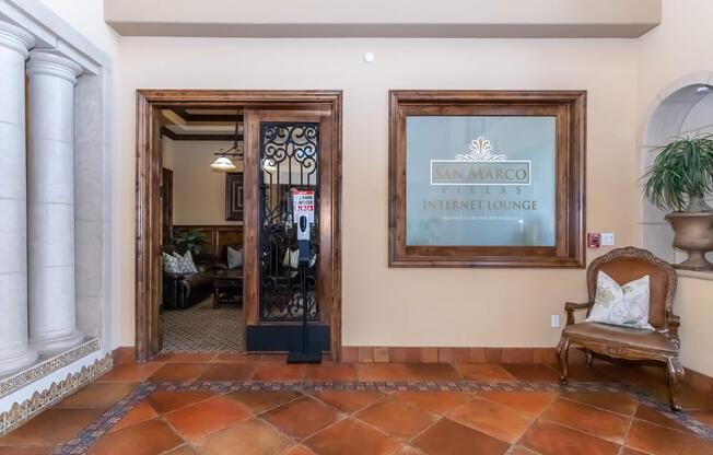 Entrance to the San Marco Villas Internet Lounge, featuring a decorative wrought iron door and a glass panel with the name. The lobby area shows tiled flooring and a comfortable seating arrangement. Pillars and a plant add to the elegant ambiance.
