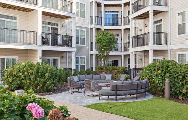 A patio with a grey couch and chairs is surrounded by a green lawn and a building with balconies.