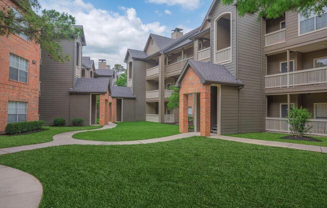 A landscaped courtyard with green grass and a walking path surrounded by two multi-story apartment buildings in a residential area. The buildings feature a mix of brick and wood siding, with balconies and large windows. The sky is partly cloudy, creating a pleasant atmosphere.