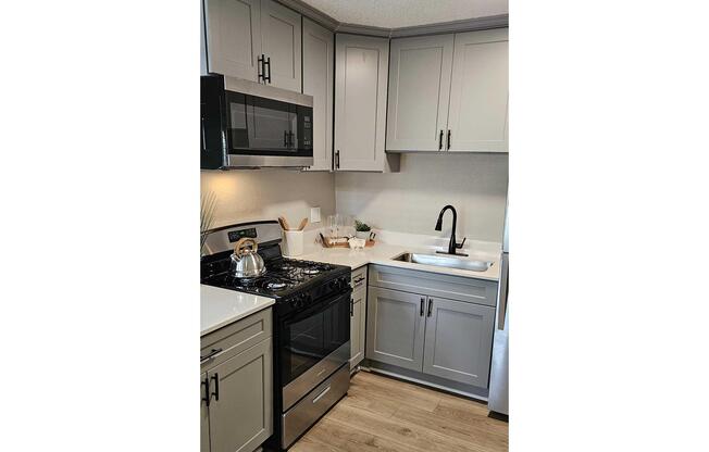 A modern kitchen featuring gray cabinetry, a stainless steel gas stove, and an integrated microwave. The sink is positioned next to a window, with minimalist decor elements on the counter, including a kettle and kitchen utensils. Natural wood flooring complements the design.