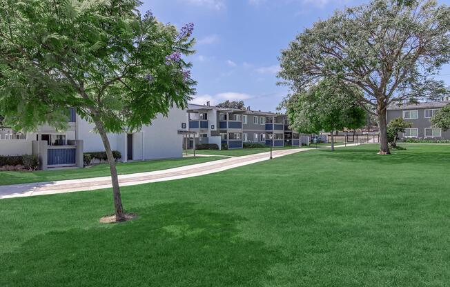 A landscaped area featuring a well-maintained grassy lawn with trees. In the background, there are modern apartment buildings and a paved walkway, creating a serene and inviting residential environment. The sky is clear with a few clouds, enhancing the peaceful atmosphere.
