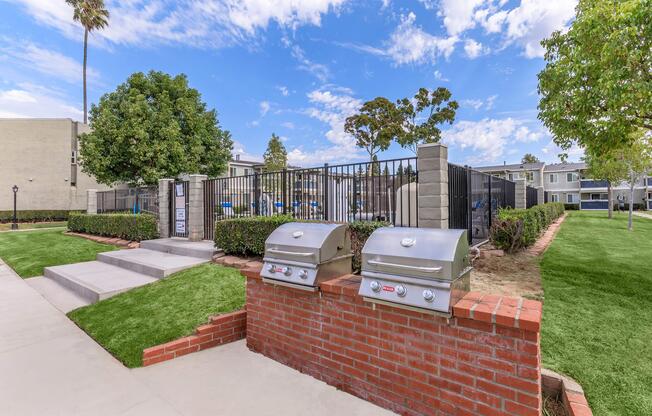 Outdoor barbecue area with two stainless steel grills situated on a brick platform. Surrounding greenery includes well-maintained grass and shrubs, with a clear blue sky and scattered clouds above. In the background, a fenced pool area and several apartment buildings are visible.