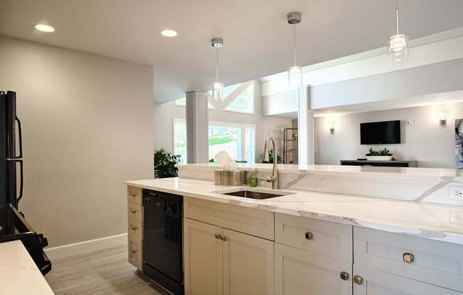 A modern clubhouse kitchen here at Andorra showcasing a spacious white island with a built-in sink, smooth countertops, light-toned cabinetry with sleek hardware, and pendant lighting overhead, opening into a bright lounge area with large windows, neutral finishes, and contemporary design details that support gatherings and resident events.