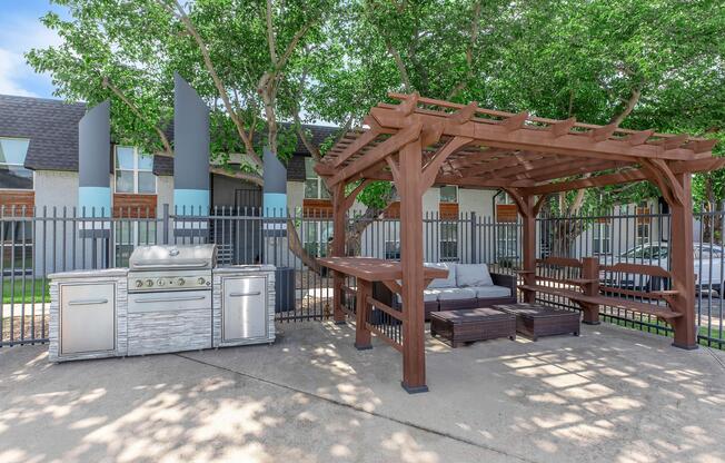 Outdoor grill area featuring a pergola with seating and a table. The space is surrounded by a fence and includes landscaping with trees providing shade. Ideal for outdoor cooking and relaxation.