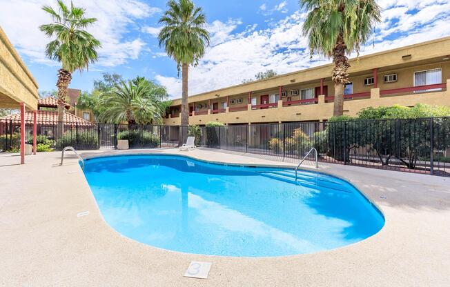 A glistening blue swimming pool surrounded by palm trees and a fence. In the background, a beige two-story building with balconies is visible under a partly cloudy sky. The pool area features a clean deck and is well-maintained, creating a relaxing atmosphere.