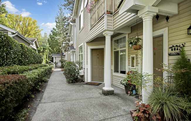 A welcoming exterior walkway here at Aspen Creek featuring a covered ground-level apartment entry with classic white columns, neutral siding, and a private front door accented by seasonal décor, hanging plants, and potted greenery, while a clean concrete path is lined with manicured shrubs, mature trees, and neighboring balconies overhead creating a residential, garden-style setting.